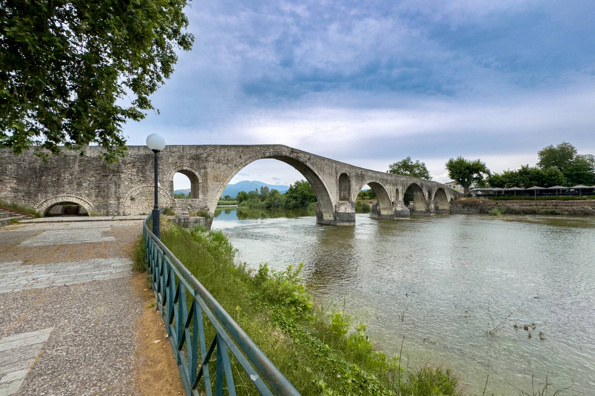The historic stone arch Bridge of Arta spanning the calm Arachthos River, framed by green trees and a paved path.