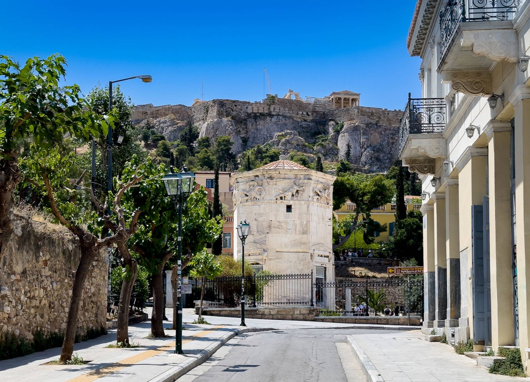 Street view in Athens, Greece, showcasing the ancient Tower of the Winds in the Roman Agora with the Acropolis and the Temple of Athena Nike visible in the background, surrounded by neoclassical architecture and lush greenery.