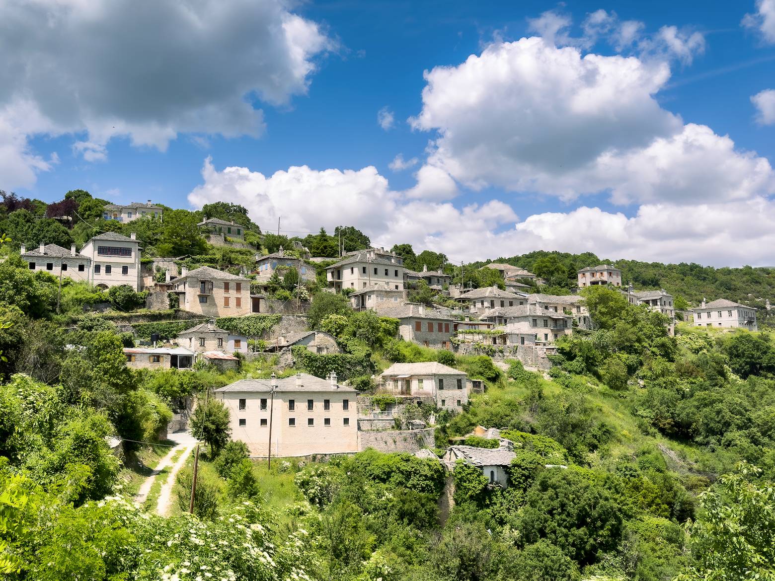 A scenic view of a traditional stone-built village nestled in the lush, green mountains of Epirus, Greece, under a blue sky with scattered white clouds.
