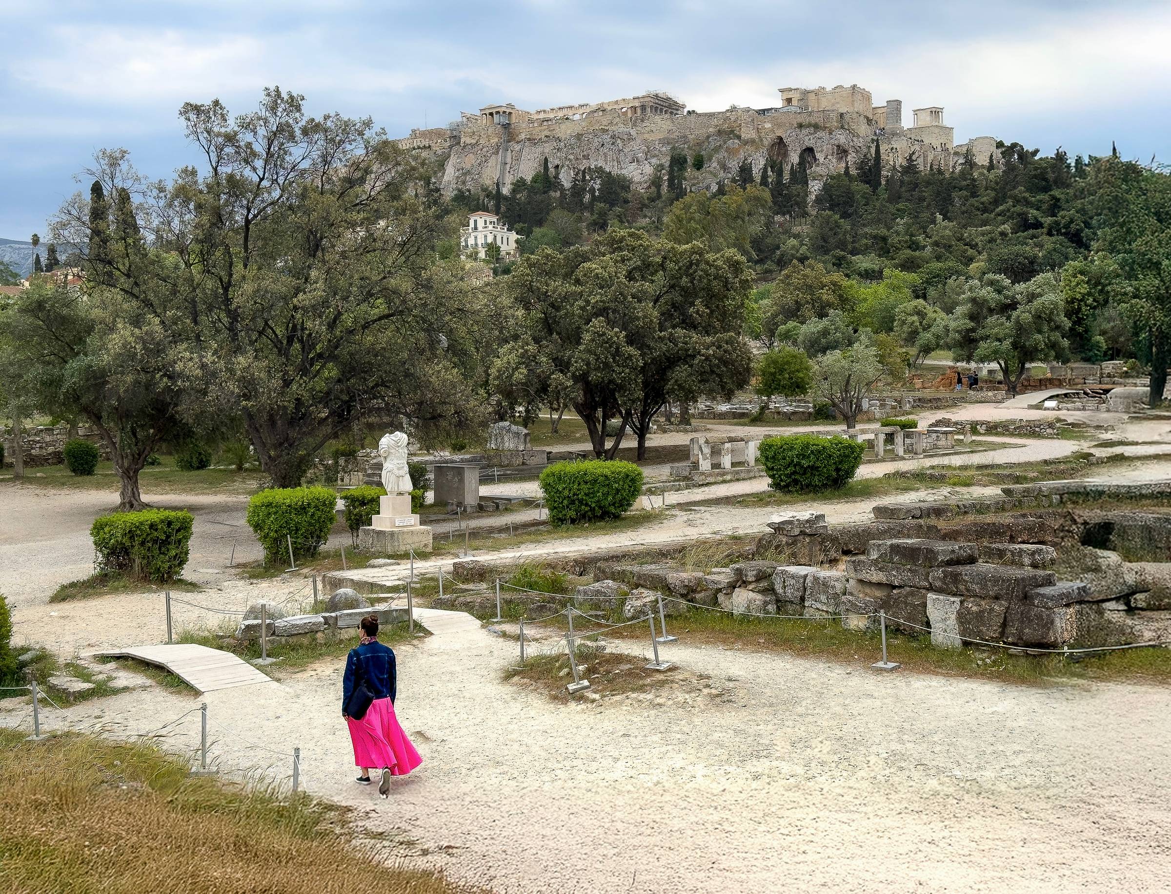 Woman in a pink skirt walking through the Ancient Agora of Athens, Greece, with views of ancient ruins, marble statues, and lush greenery, set against the backdrop of the iconic Acropolis hill and Parthenon.