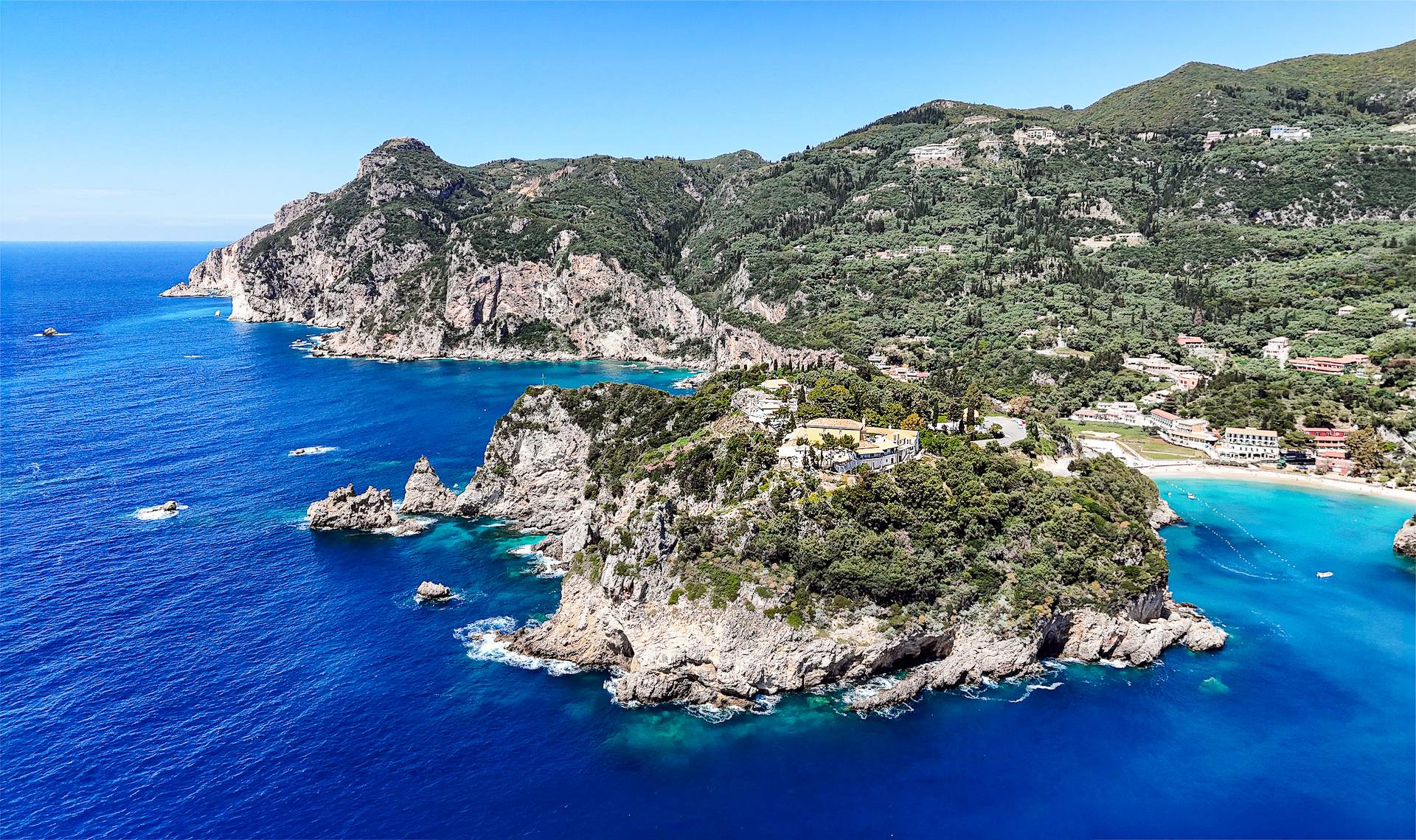 Aerial panoramic view of Palaiokastritsa, Corfu, showing rugged green-covered cliffs, turquoise bays, rocky coves, and scattered buildings along the hillside and shoreline beneath a clear blue sky.