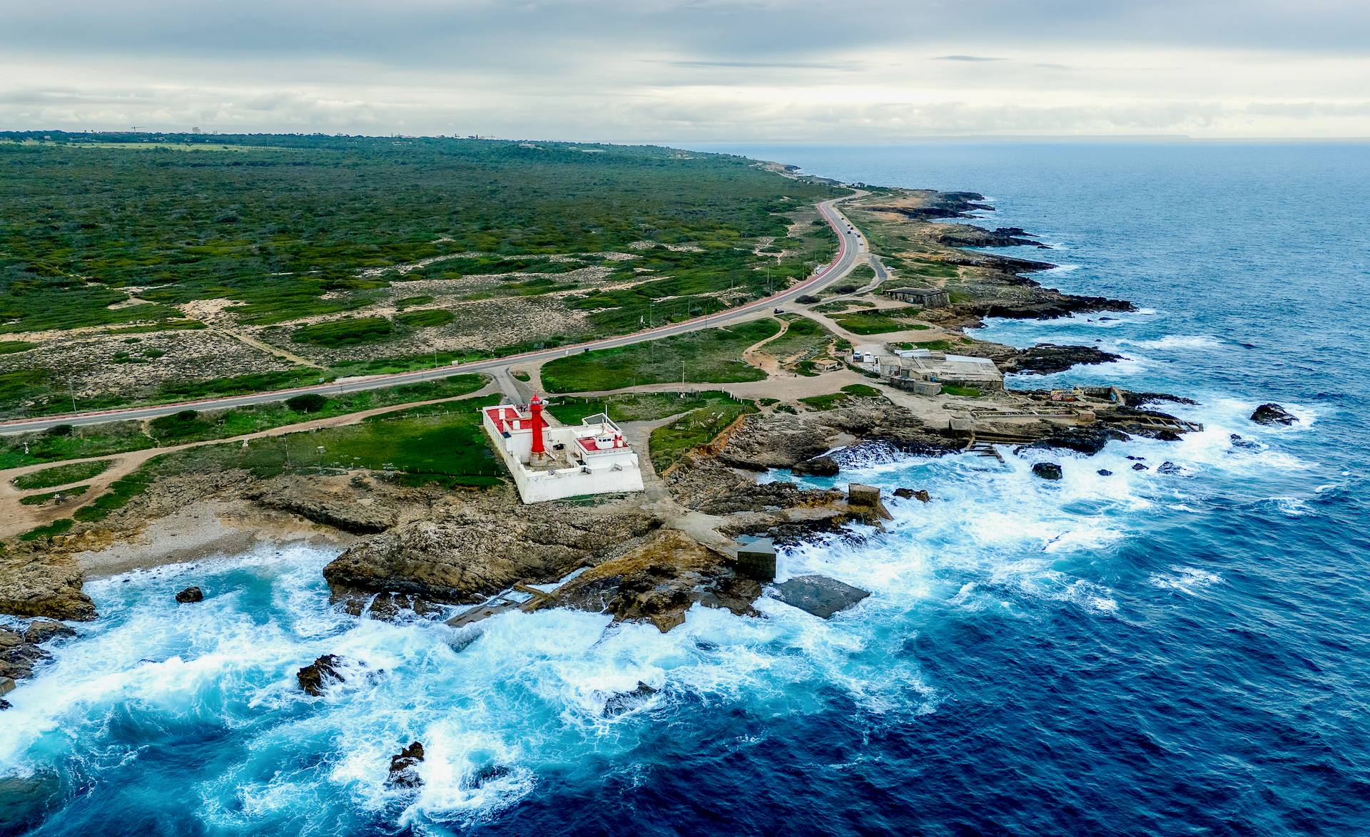 Aerial view of a white lighthouse with a red roof on a rocky coastline, featuring crashing blue waves and a coastal road under a cloudy sky.