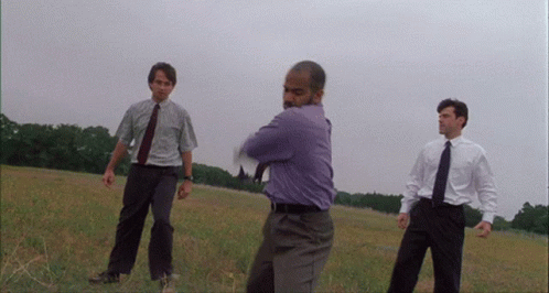 A scene from the movie Office Space where three men is business attire destroy a printer in a field with a baseball bat