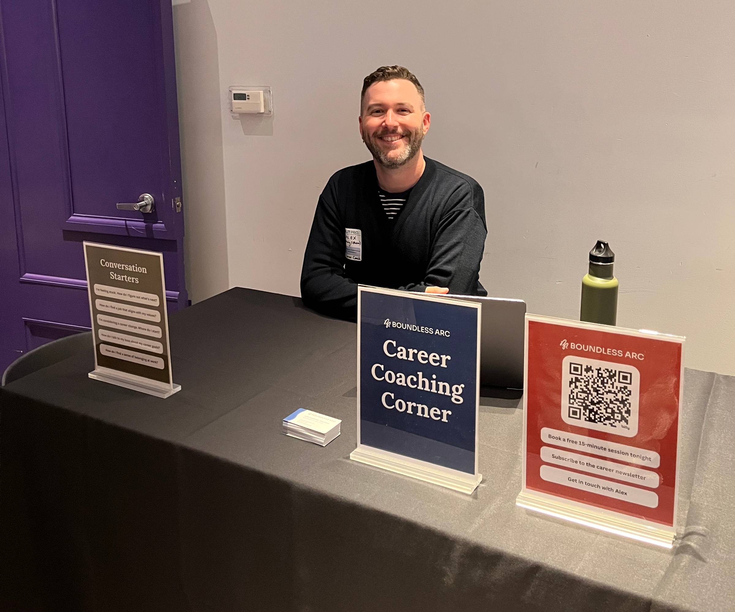 Alex is sitting behind a table at a networking event with the sign "Career Coaching Corner" and smiling at the camera.