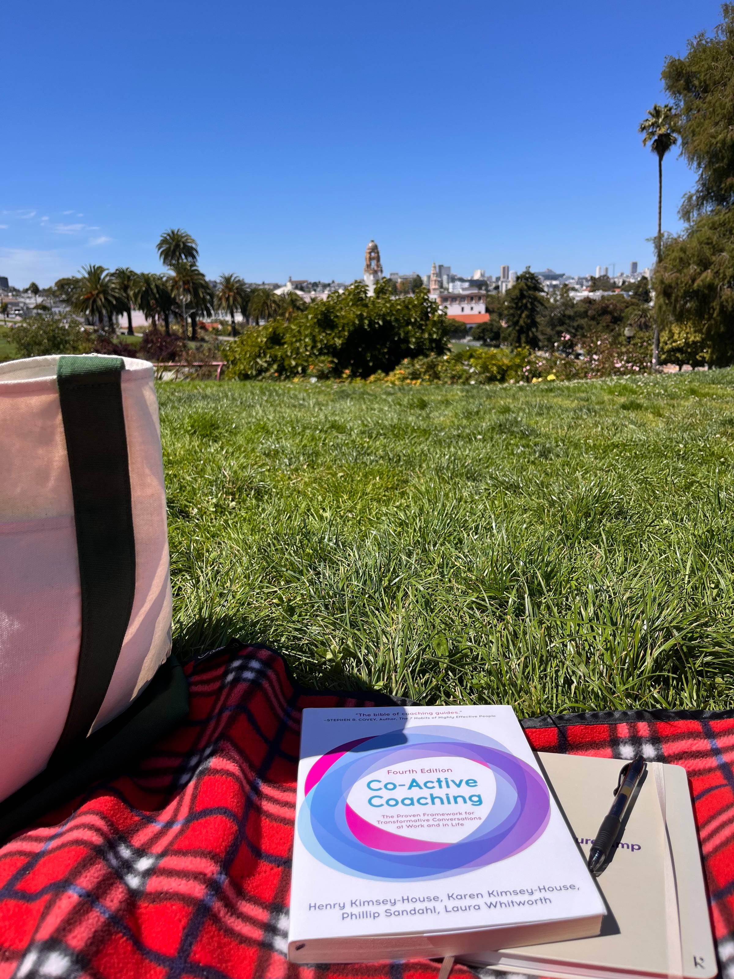 A copy of 'Co-Active Coaching' rests on a red plaid blanket in Dolores Park, San Francisco, next to a notebook, pen, and tote bag, with a grassy field and the city skyline in the background.
