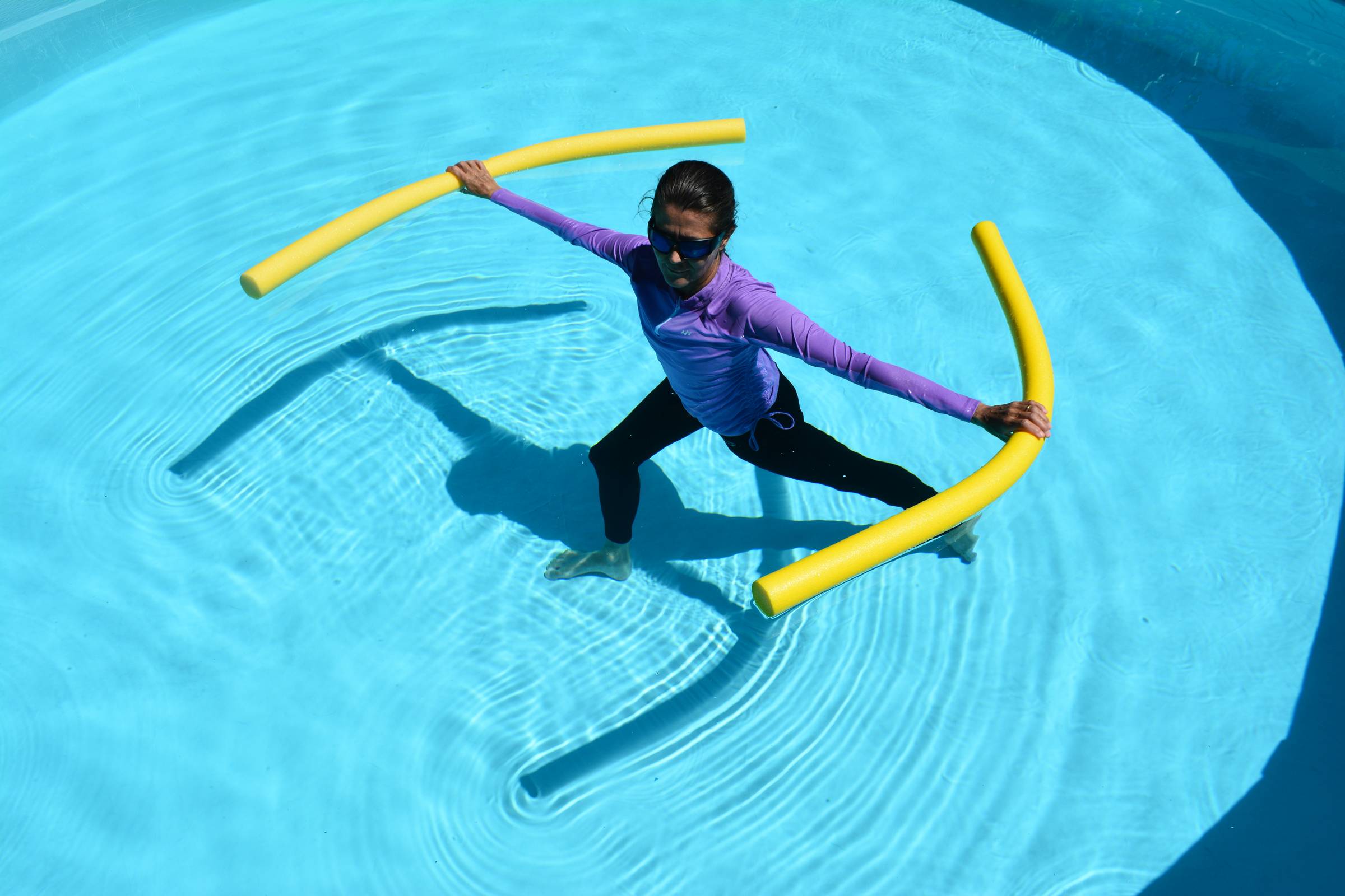 Christa Fairbrother in the pool doing an aqua yoga Warrior I spinal twist.  She's wearing black swim pants, a lavendar swim top and sunglasses.  In her hands which are spread open on the surface of the water are two yellow pool noodles.