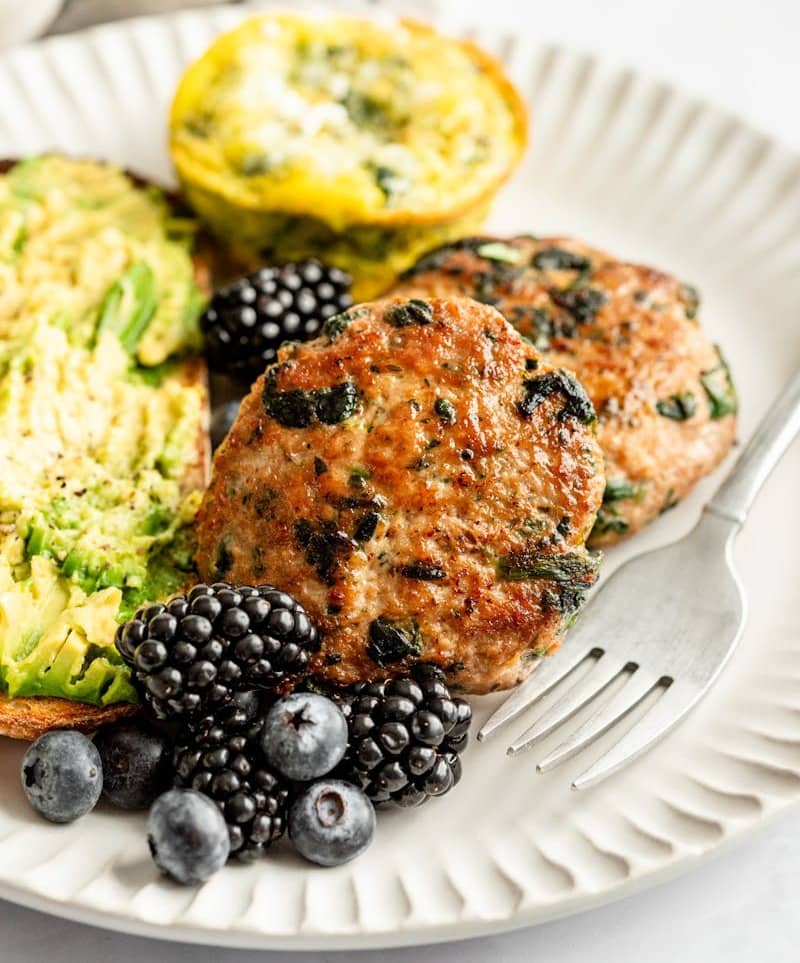 Egg bites, aveggie-loaded sausage, avocado toast, and fresh blueberries and blackberries next to a fork on a plate.