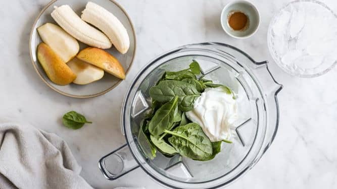 Spinach and yogurt in a blender cup sitting on a counter. The blender cup is surrounded by additional smoothie ingredients such as a cup of ice, a plate of bananas and apples, and a small container with spices.