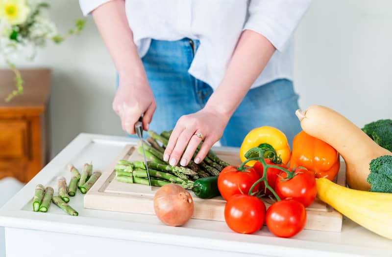 Asparagus being chopped on a cutting board. On the cutting board and on the countertop surrounding the board are whole tomatoes, bell peppers, a summer squash, broccoli, a butternut squash, and an onion.