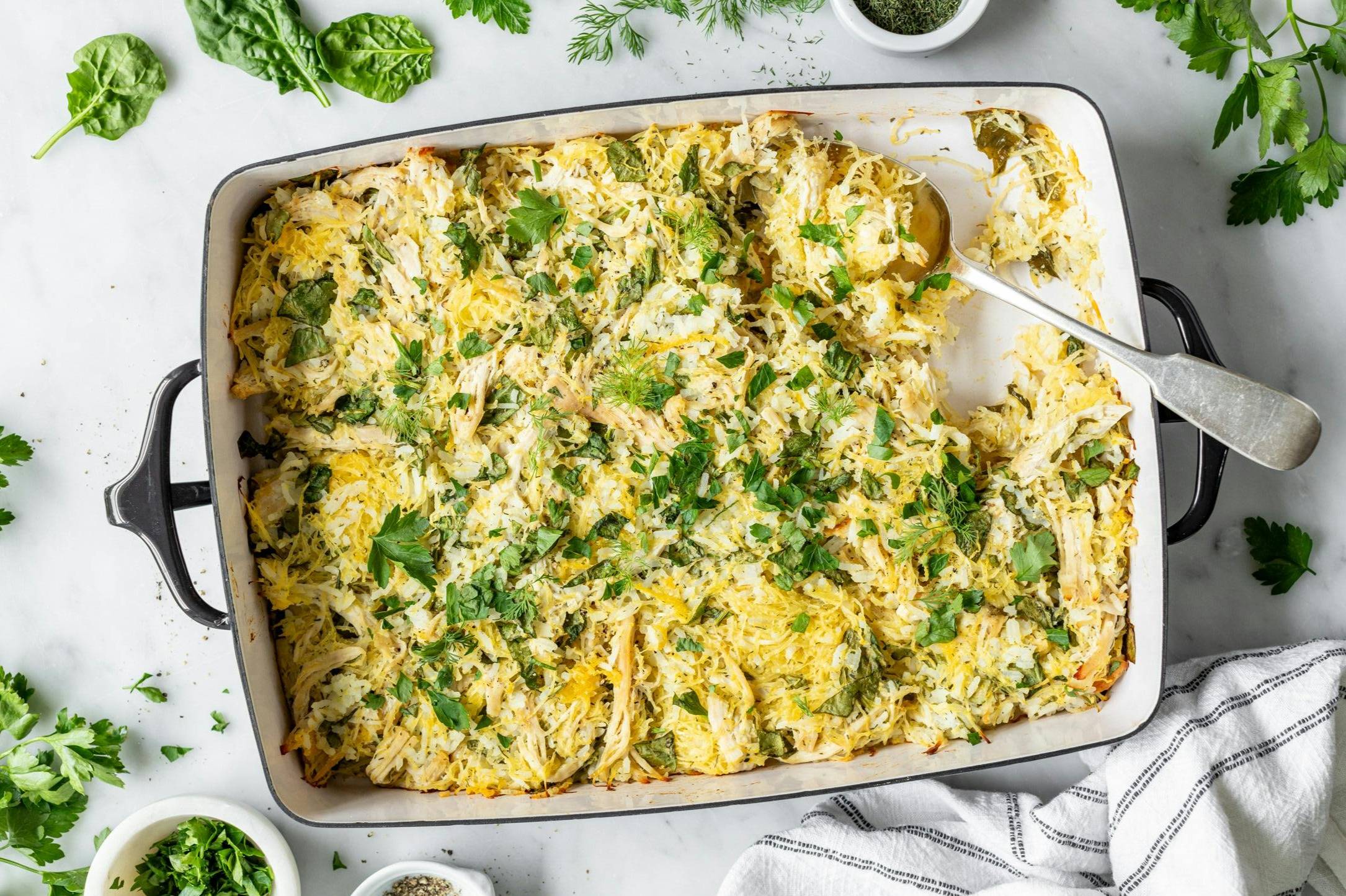 A veggie-loaded casserole in a casserole dish on a countertop. The dish is surrounded by a cloth napkin, and some fresh herbs and spinach leaves strewn about the countertop.