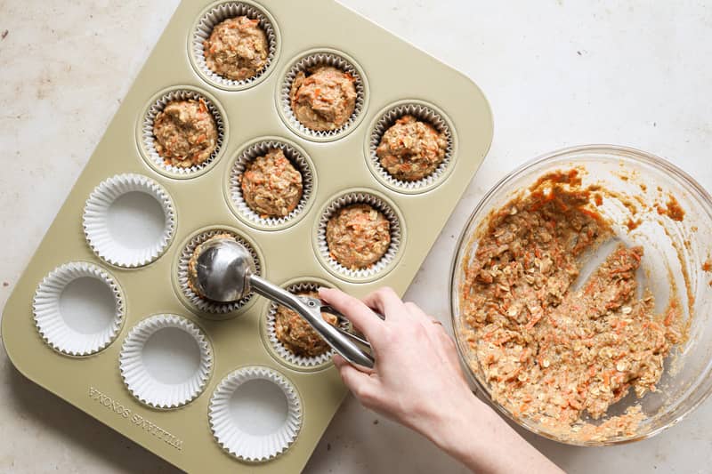 Muffin batter being scooped with a cookie scoop from a glass bowl into a muffin tin filled with paper liners