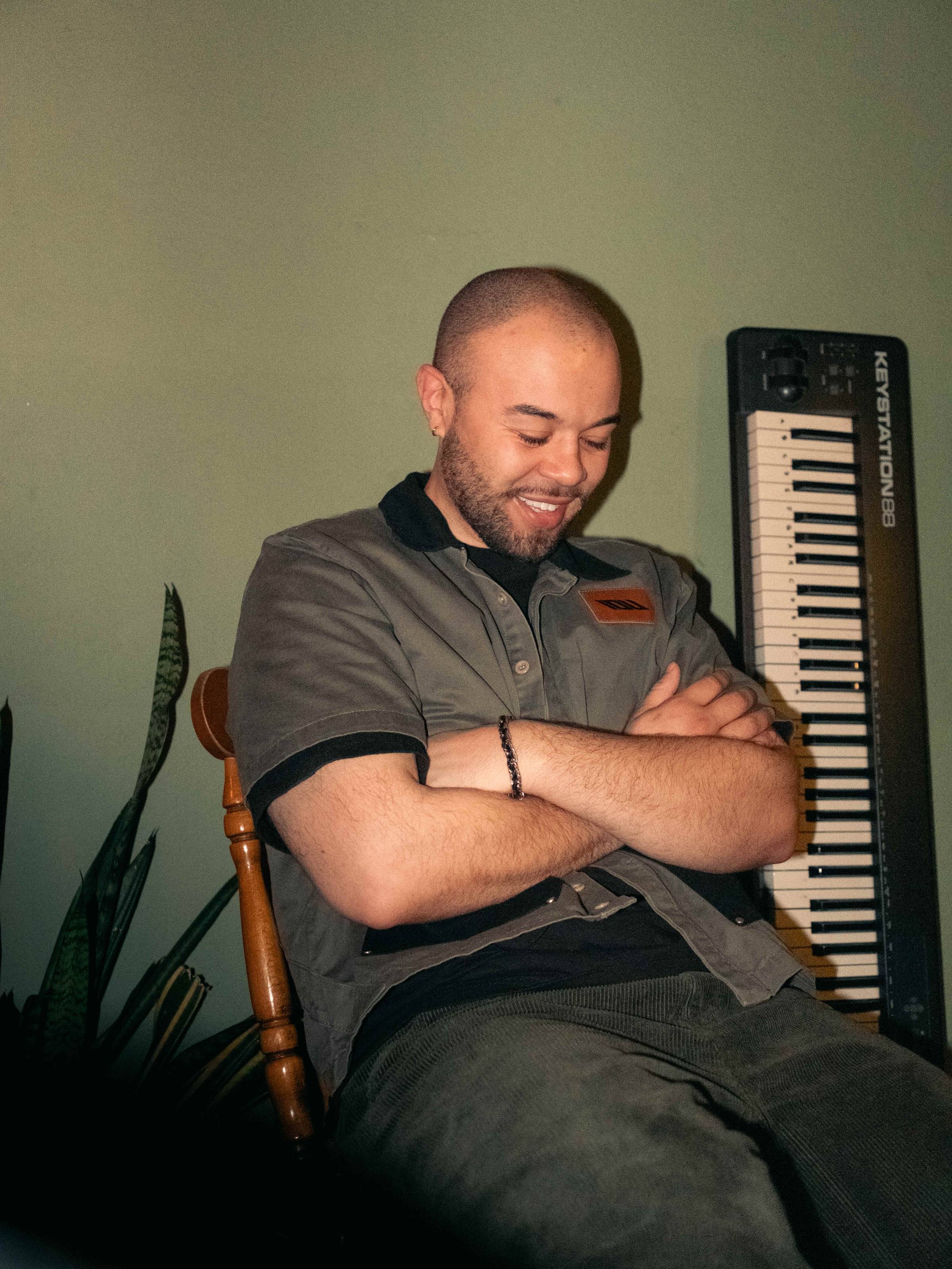 a light skinned Black man is smiling while sitting on a wooden chair inside with a keyboard behind him