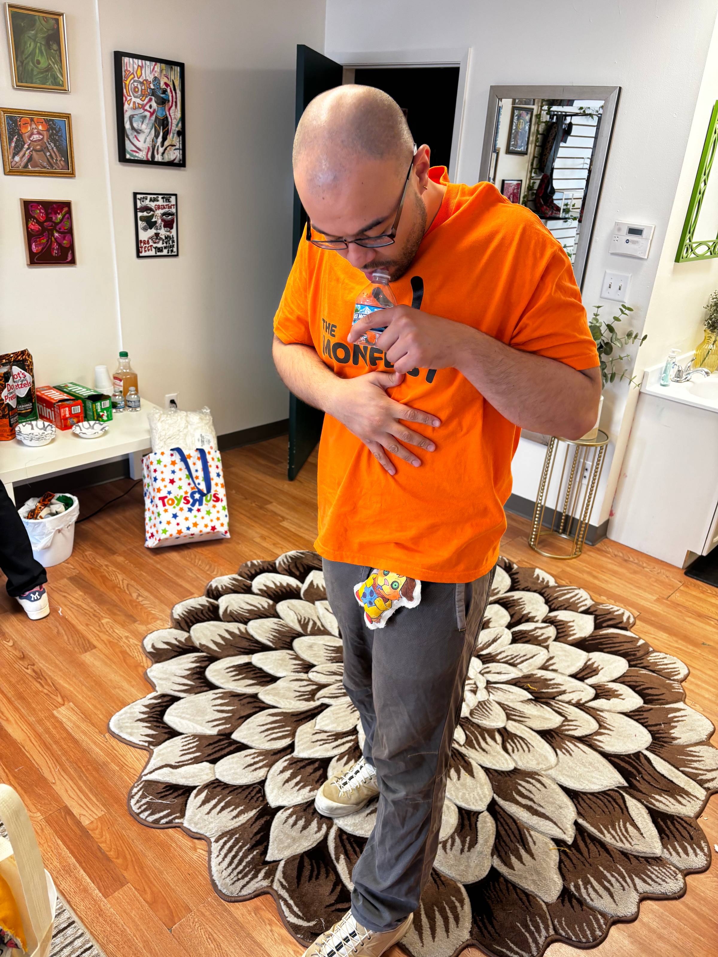 Kub stands in a bright orange monfest shirt looking down at a plushie attached to a belt loop