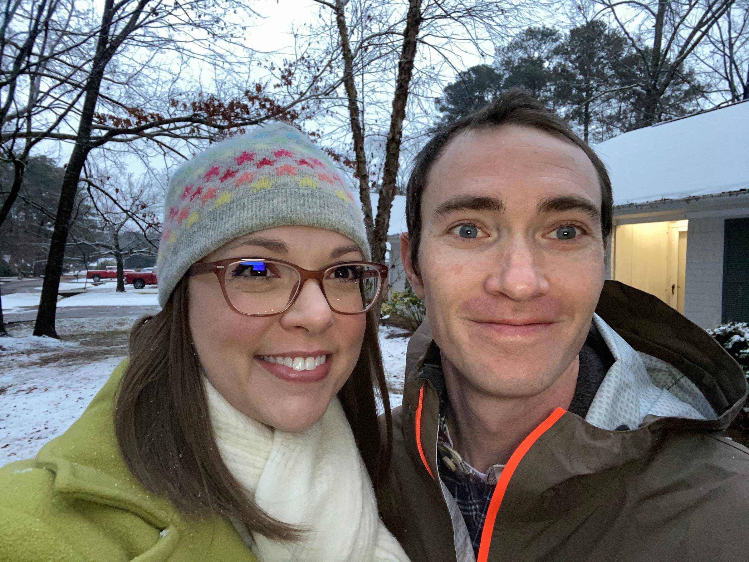 Two people standing outside with snow on the house and trees in the background. One person is a white female with medium-length brown hair and glasses. She is smiling and wearing a grey knit cap, a lime green coat, and a cream colored scarf. To her right 