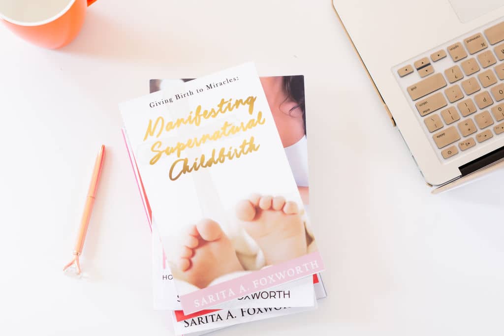 A stack of books is placed on a white surface next to a laptop and an orange cup. The top book is titled "Manifesting Supernatural Childbirth" by Sarita A. Foxworth, featuring a cover image of baby feet. A pink pen is positioned beside the books.