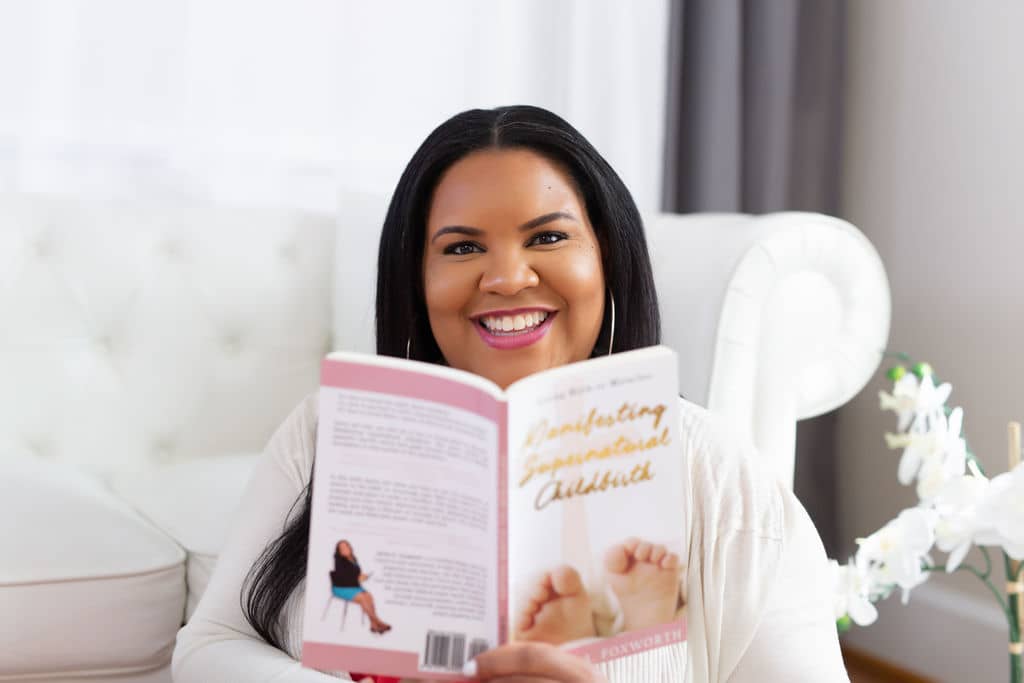 A woman with long dark hair is sitting on a white couch, smiling while holding a book titled "Manifesting Supernatural Childbirth." The room is bright, with light-colored furniture and decor, including a white flower arrangement nearby.