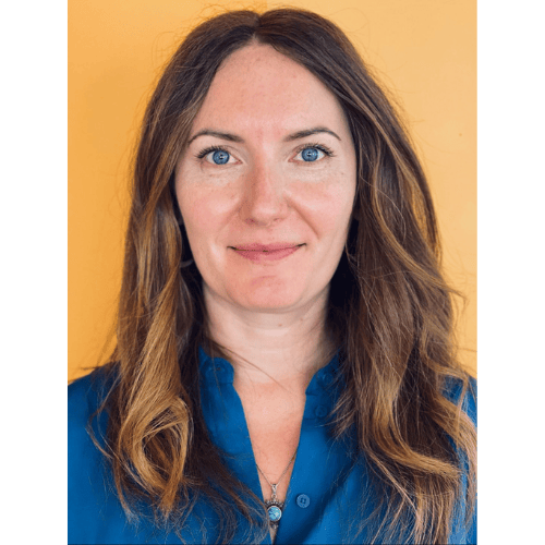 A person with long wavy brown hair is wearing a blue shirt and looking directly at the camera. The background is a solid orange color.