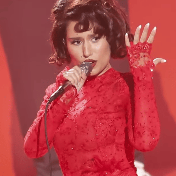 Woman wearing bright red dress sings and holds up her hand showing where a wedding ring would be, if she were married