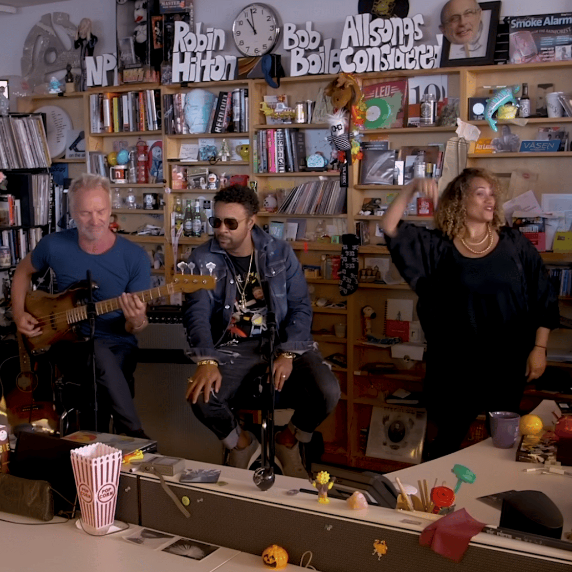 Sting, Shaddy, and others sing and play instruments in front of overflowing bookshelves