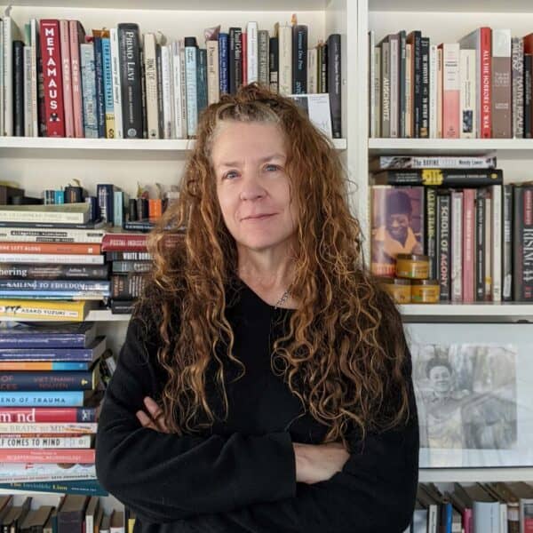 Headshot - woman with long, curly, brown hair stands in front of bookshelves