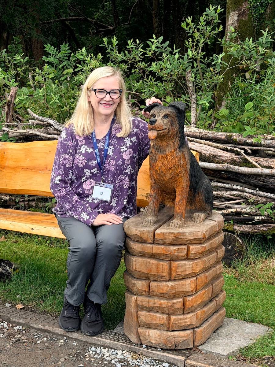 Bonni sits on a bench "petting" a dog that is carved into the edge of the bench
