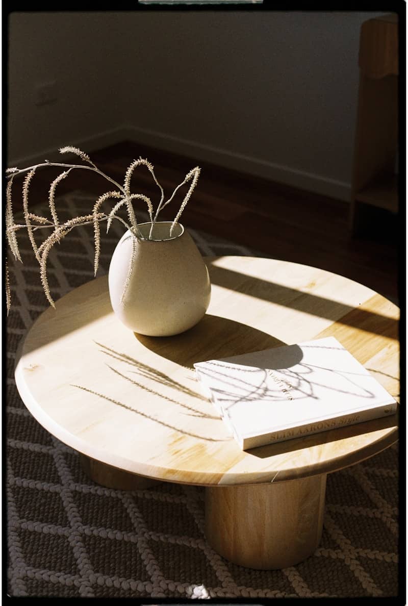 A vase and book sit on a sunlit coffee table.