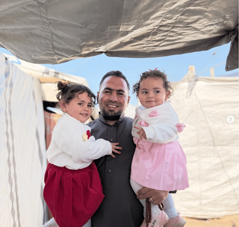 Basel holds his daughters Lara and Mira as they stand outside their tent