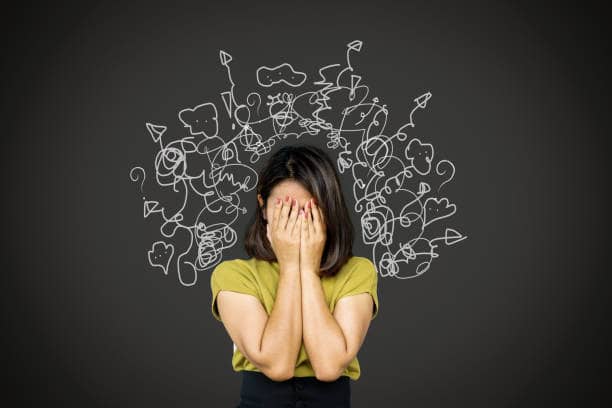 A photo of a woman with her hands covering her face. Behind her on a black wall is white pencil drawing depicting a mind of chaos... arrows curving an d pointing every which way, cartoon voice bubbles and question marks everywhere. The drawing appears to come from the woman's mind and spiral outward from there.