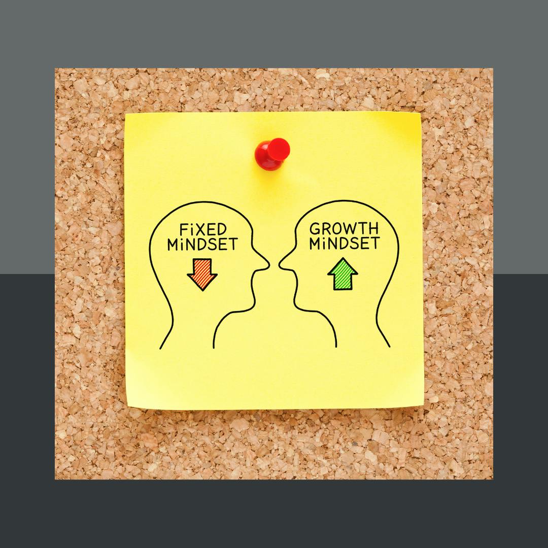 A corkboard shows a post-it note held up with a red pushpin. On the post-it are two images, outlines of a human head. The heads are facing each other. The first has the text "Fixed Mindset" with a red arrow pointing down in the middle of it. The second has the text "Growth Mindset" with a green arrow pointing up inside it.