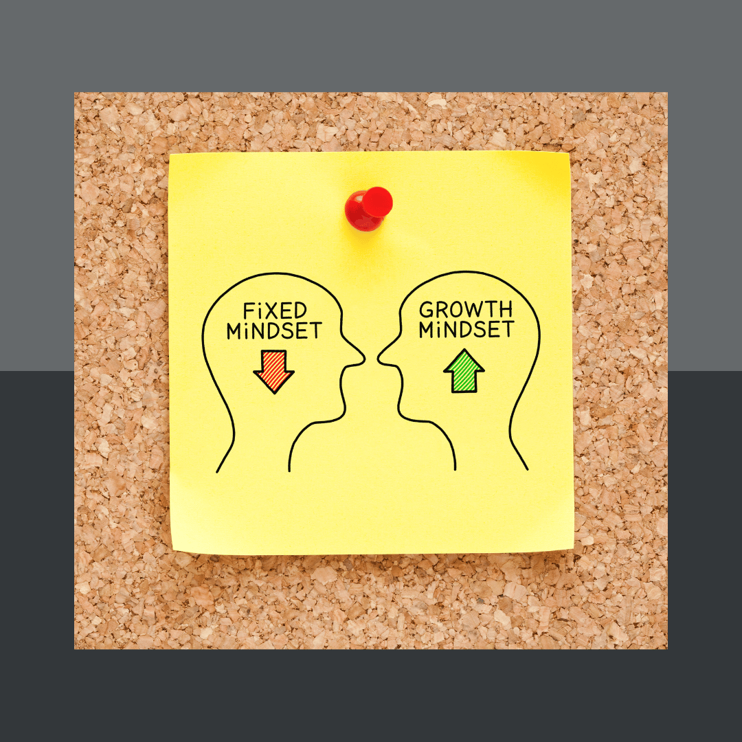 A corkboard shows a post-it note held up with a red pushpin. On the post-it are two images, outlines of a human head. The heads are facing each other. The first has the text "Fixed Mindset" with a red arrow pointing down in the middle of it. The second has the text "Growth Mindset" with a green arrow pointing up inside it.