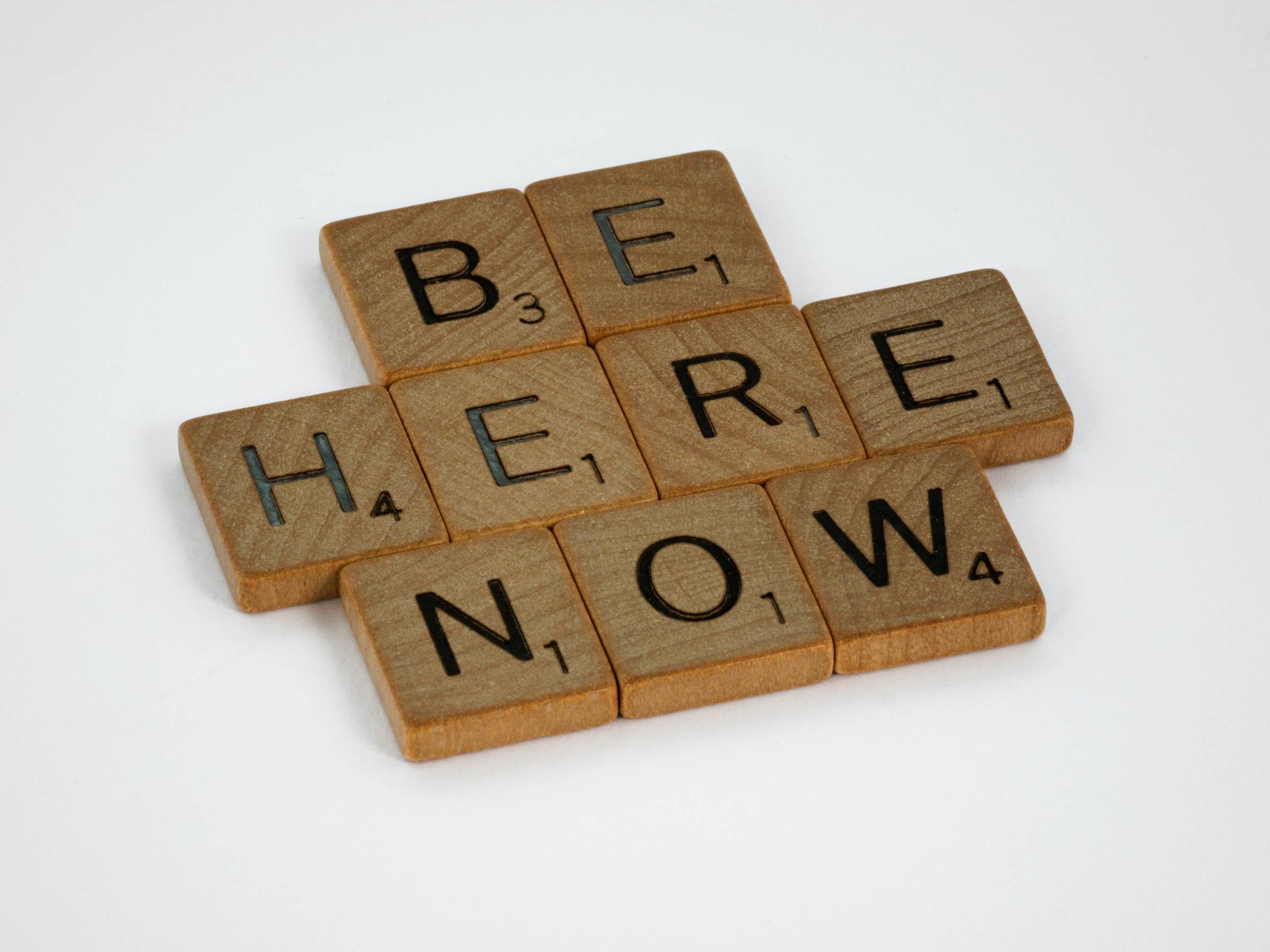 Scrabble letter tiles spell out the words "Be Here Now".