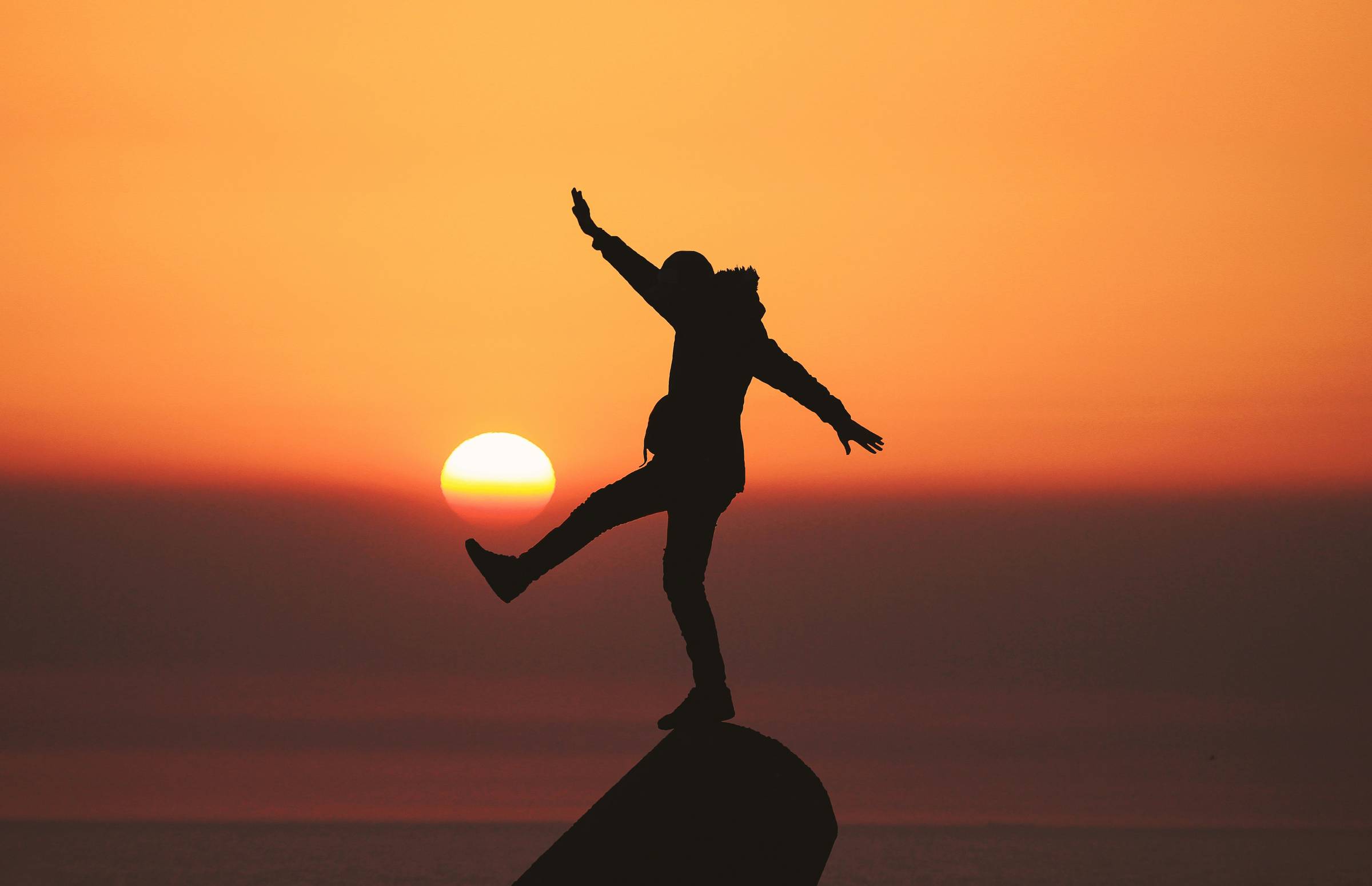 A silhouette of a person balancing on one foot standing atop a large rock against a sunset backdrop 