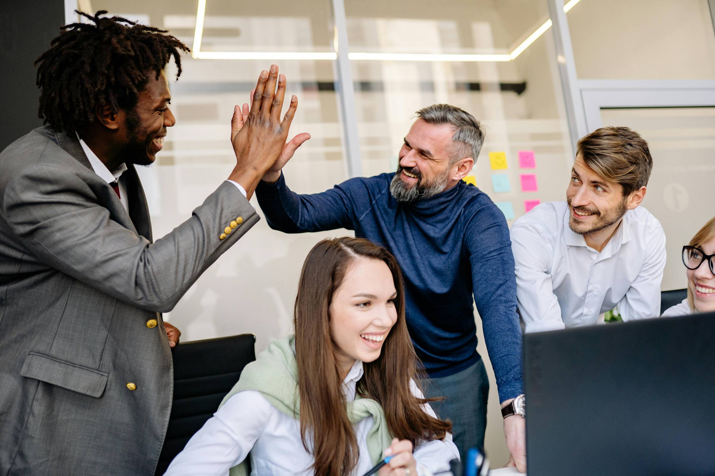 A photo of five employees around an open laptop. Two of them are giving each other a high five while the others look on and smile.