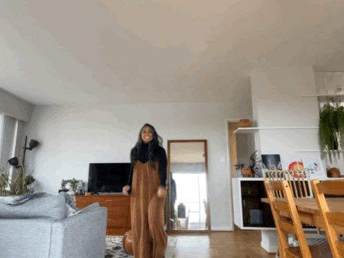 A gif of brown woman in orange overalls and a black turtleneck. She's running up to the camera and taps her heels together in the air while leaning on the side of a couch for support with a giant smile on her face.