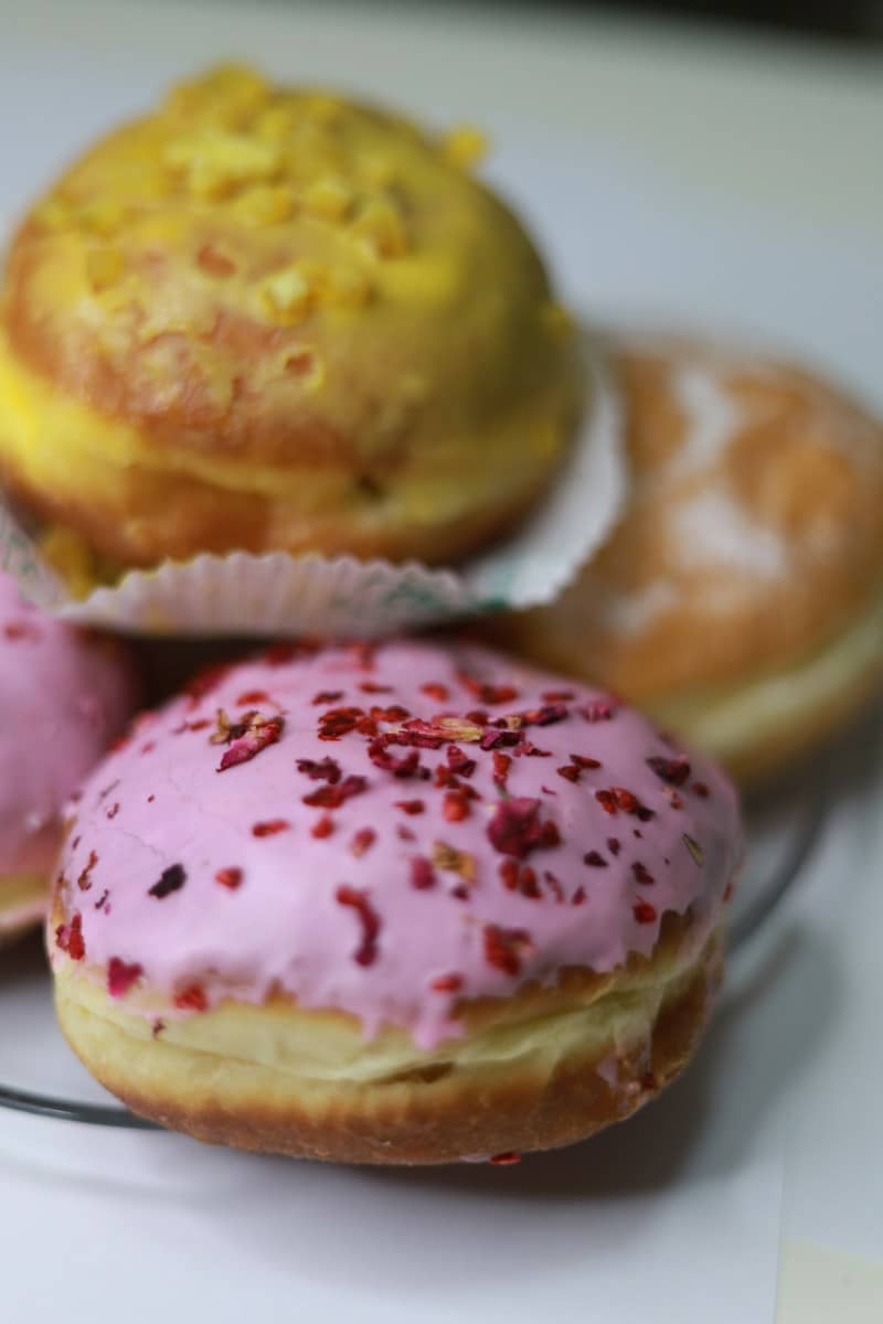 A pink glazed donut topped with dried flower petals beside yellow and glazed donuts