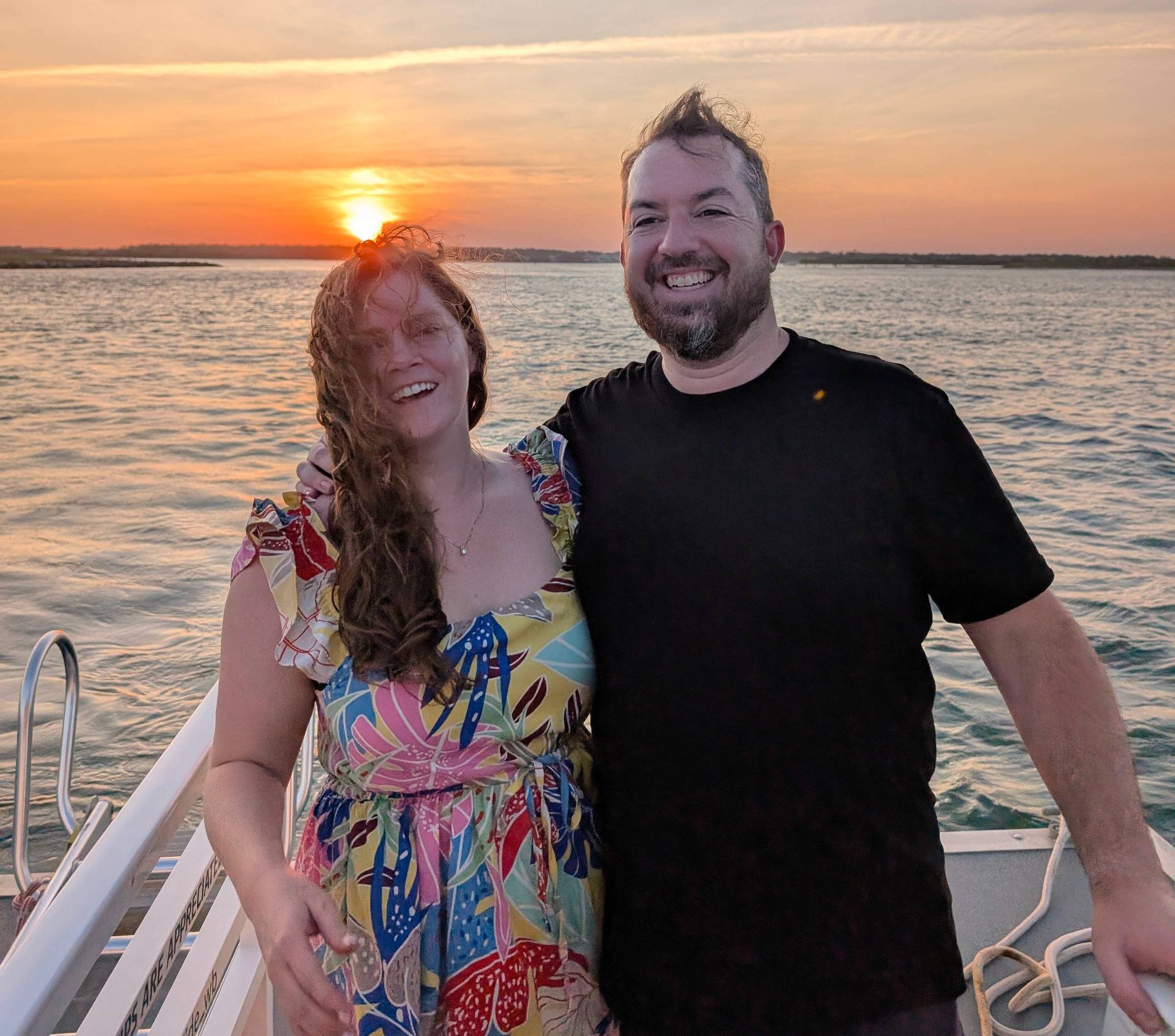 Natalie and Mike standing on the front of a boat with the sunset behind them. It is windy out so natalie's hair is covering her face and Mike's hair is blowing upward. They are smiling and laughing