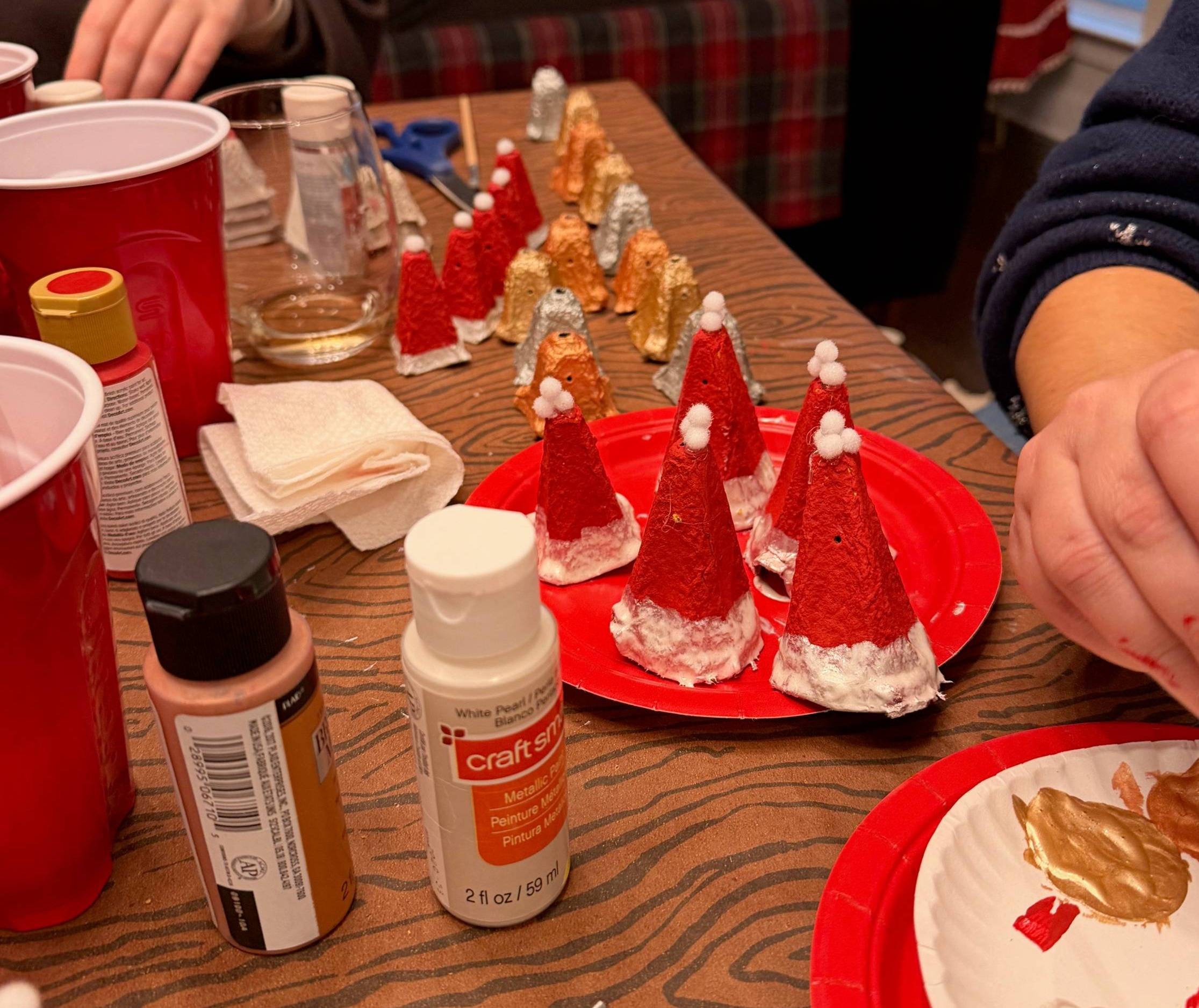 A table covered in crafting supplies. Small santa hats and bells are being made out of cardboard egg cartons