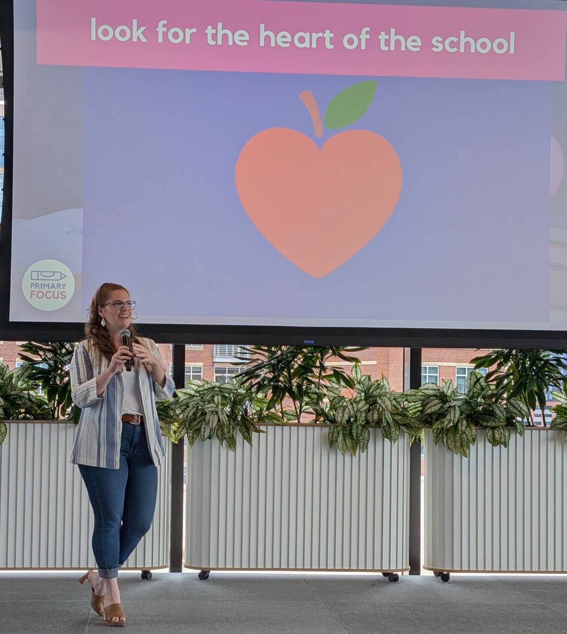 Natalie standing on stage holding a microphone. The slides behind her say "look for the heart of the school"