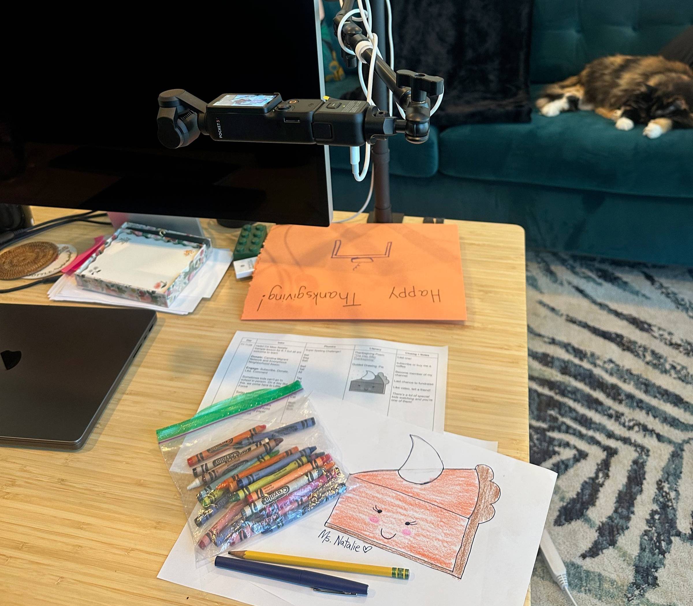 A picture of Natalie's desk with a hand drawn picture of a slice of pie, overhead camera, and cat napping on the couch in the background