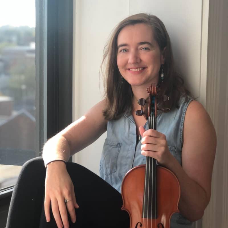 Hannah is smiling and holding her fiddle in an urban apartment setting