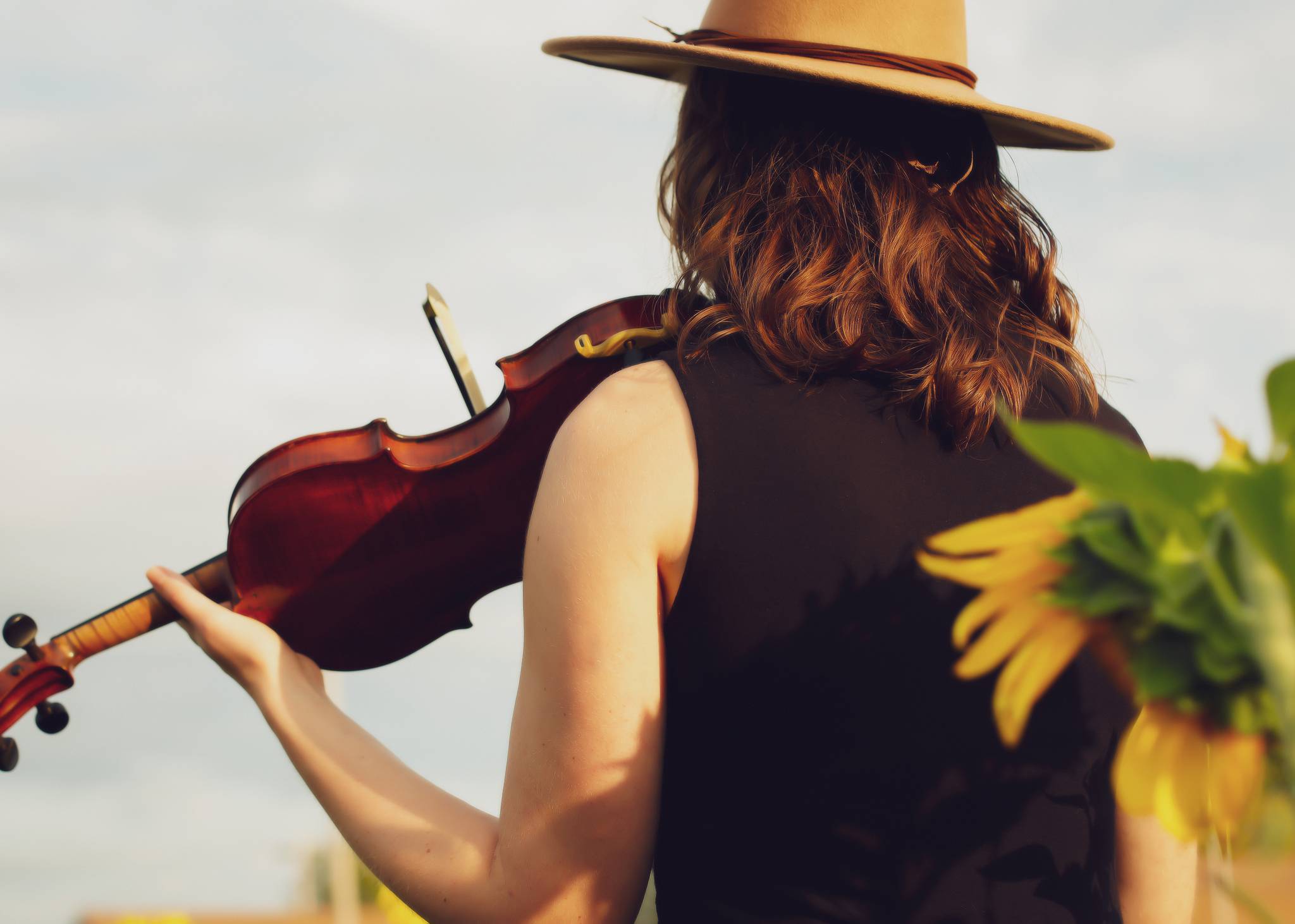 A girl wearing a western hat holding a fiddle in a field of sunflowers