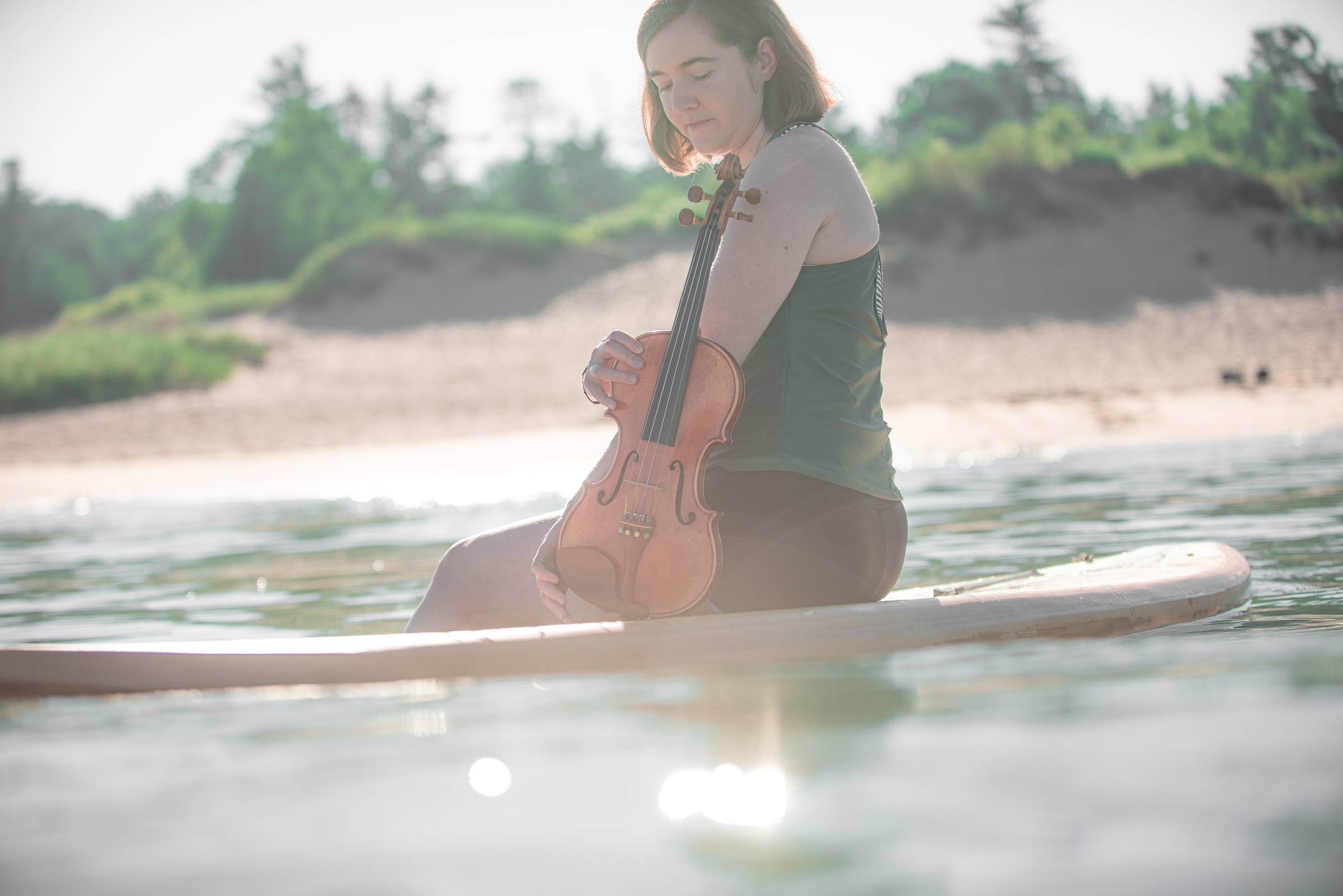 Hannah floats near the shores of Lake Michigan on a paddleboard holding her fiddle.