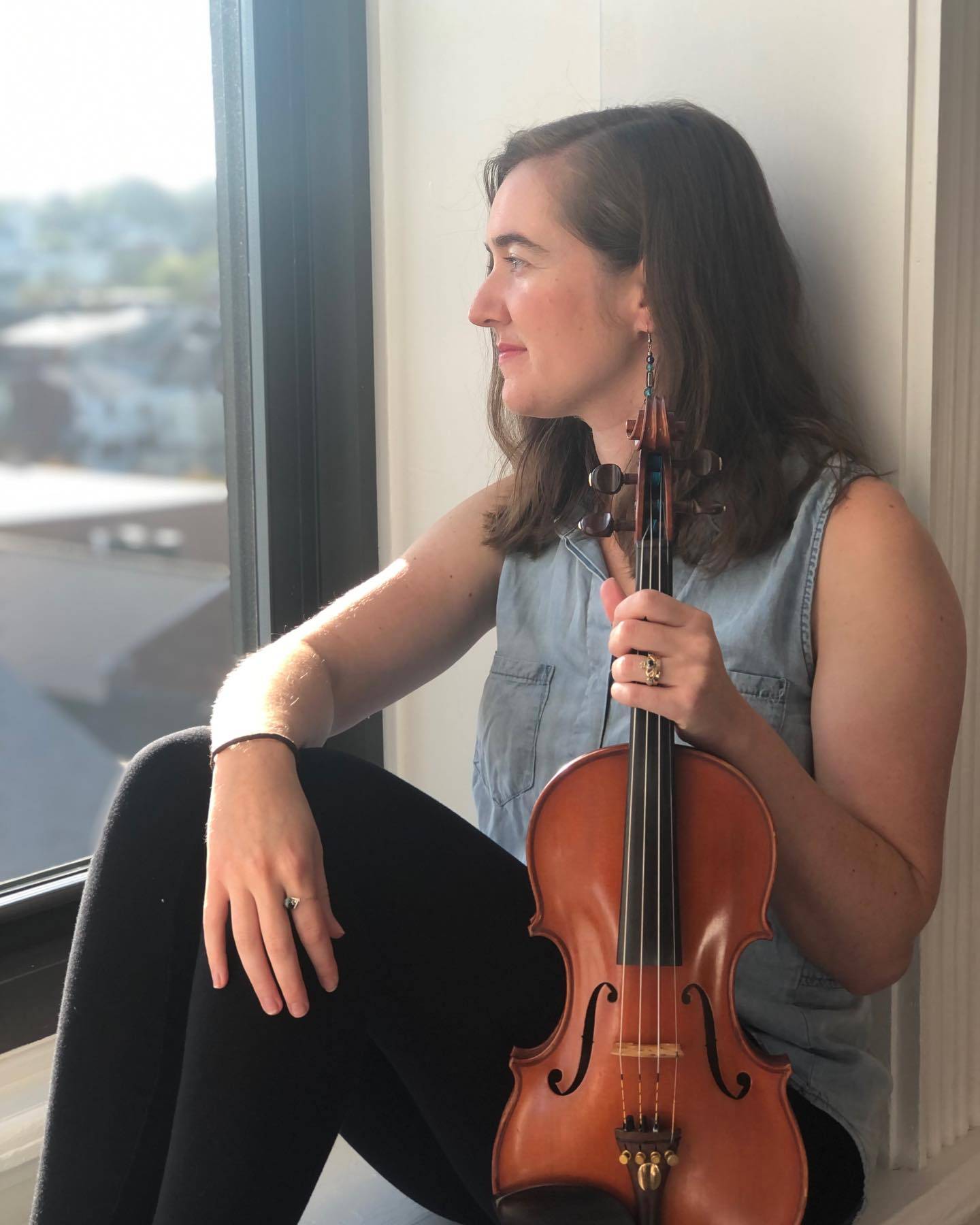 Hannah sits with her fiddle in an urban apartment looking out at the city