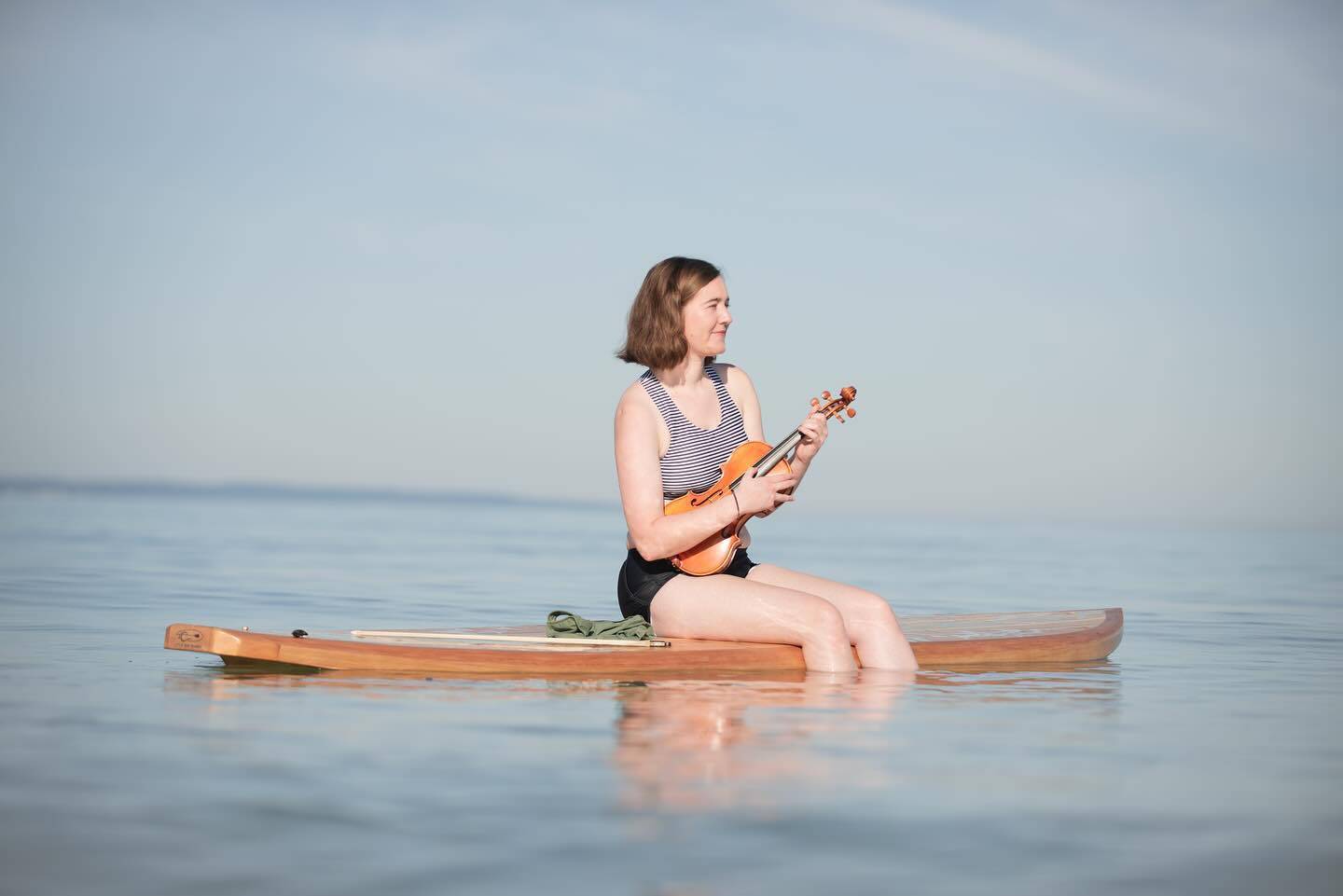 Hannah sits on a paddle board with her fiddle on Lake Michigan