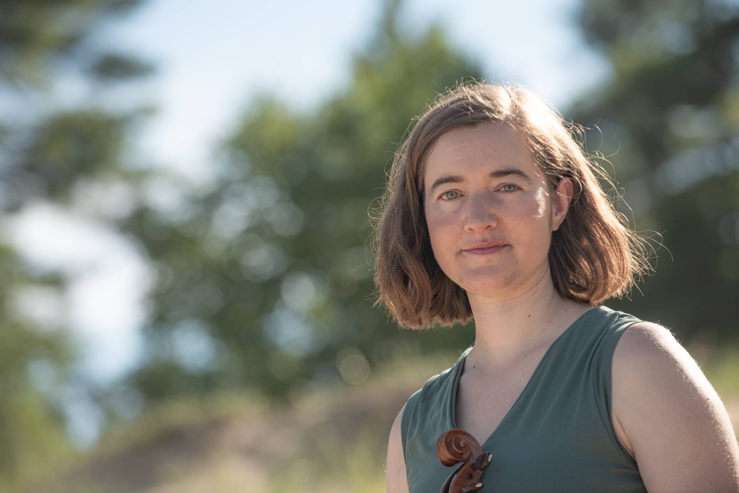 A girl in a green dress holding a fiddle outside with a blue sky and trees in the background