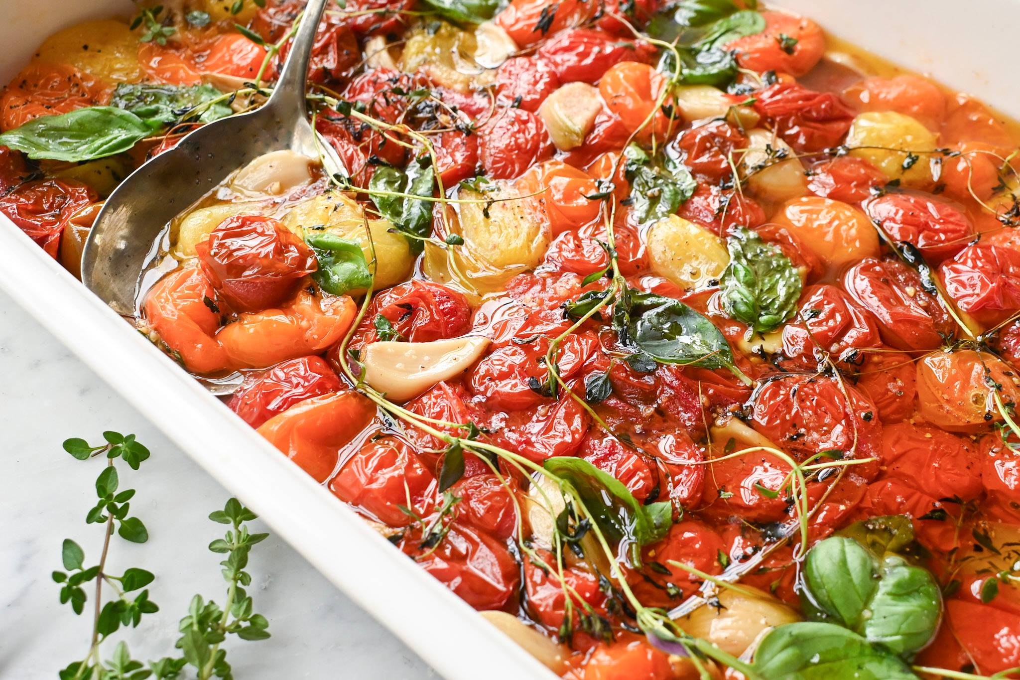 Cherry tomato confit with garlic, thyme, basil, and olive oil in a white baking dish.