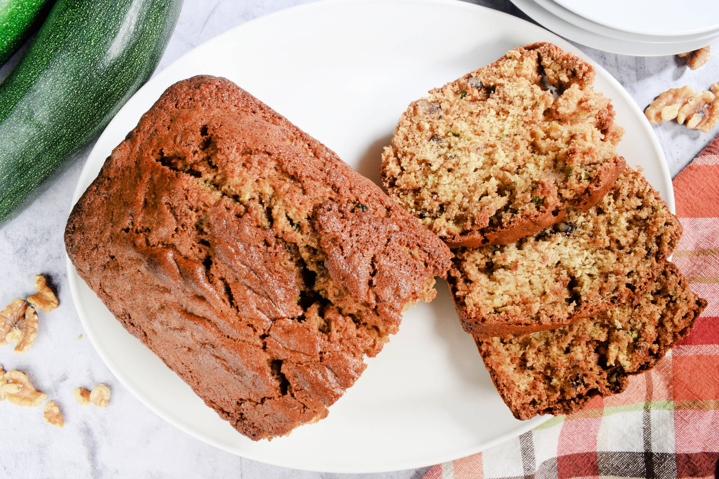 Sliced sourdough zucchini bread on a white plate with fresh zucchini and walnuts nearby.