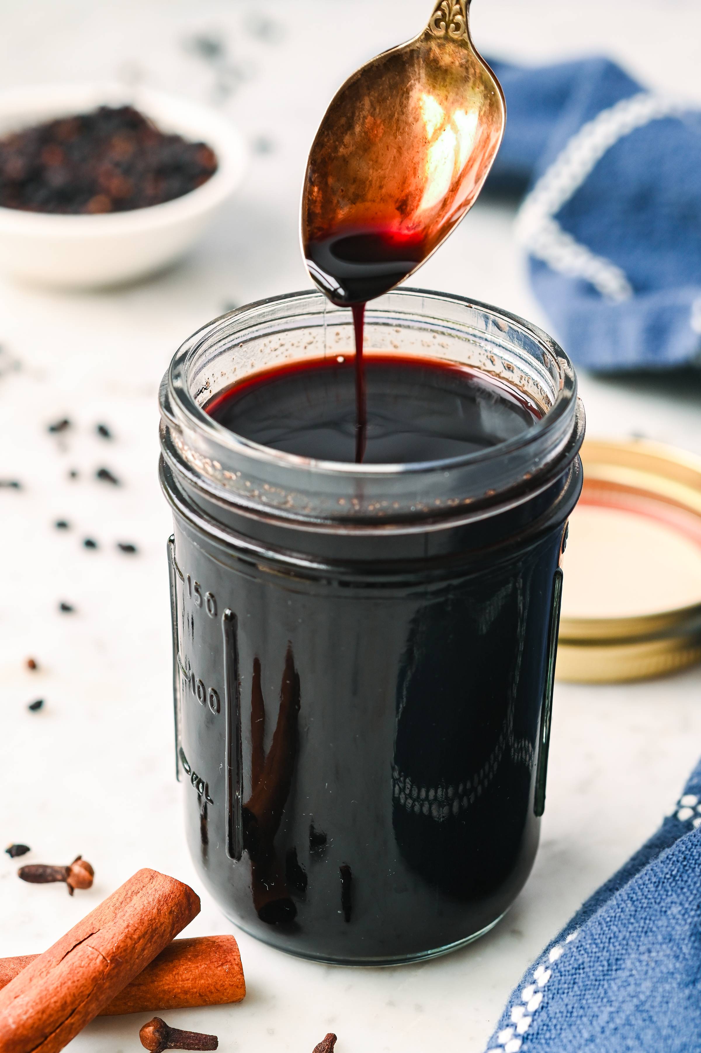 Homemade elderberry syrup dripping from a spoon into a mason jar with cinnamon sticks, cloves, and dried elderberries nearby.
