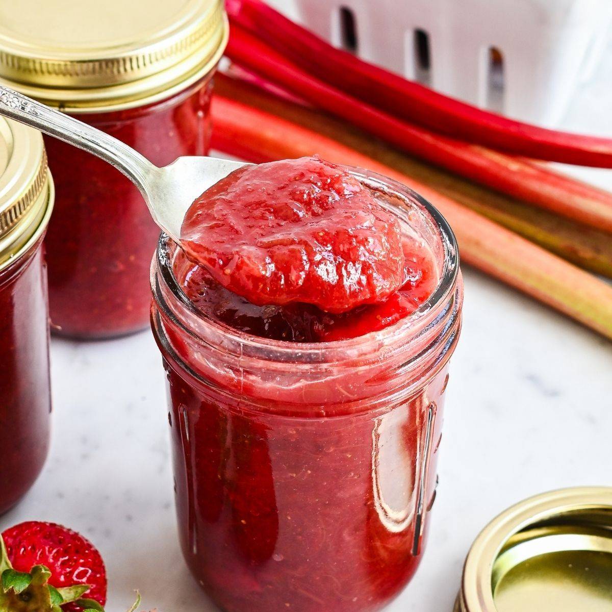 Jar of homemade strawberry rhubarb jam with a spoonful of jam, fresh rhubarb stalks, and strawberries nearby.
