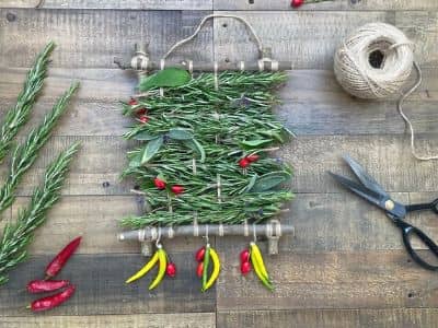 A small woven wall hanging made from fresh rosemary, bay leaves and mixed herbs, decorated with hanging chillies in red and yellow, displayed on a weathered wooden surface with a ball of twine and scissors alongside.